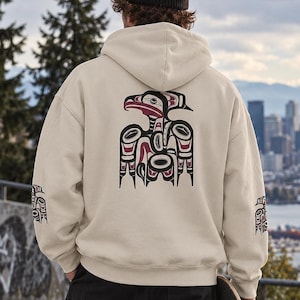 Op de afbeelding: Een beige hoodie met een zwart, wit en bordeauxrood grafisch ontwerp van een gestileerde vogel op de rug en kleinere bijpassende vogelafbeeldingen op elke mouw. De hoodie wordt gedragen met een zwarte muts en een zwarte broek.