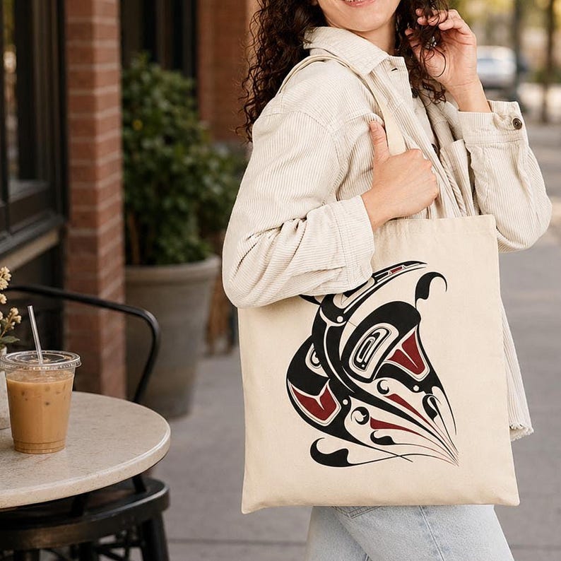 AI-generated lifestyle mockup of a natural cotton canvas tote bag with original Pacific Northwest Coast inspired raven formline artwork, worn with a cream jacket and light denim beside a cozy city café with iced coffee, brick storefronts, and soft summer-to-fall light.