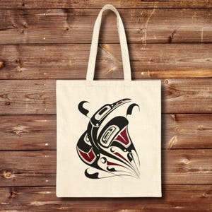 Natural cotton canvas tote bag featuring a bold Northwest Coast–inspired raven totem design in black and deep red, centered on the front. The bag hangs against a warm wooden plank wall background, showing long reinforced shoulder straps and a structured flat-bottom shape.
