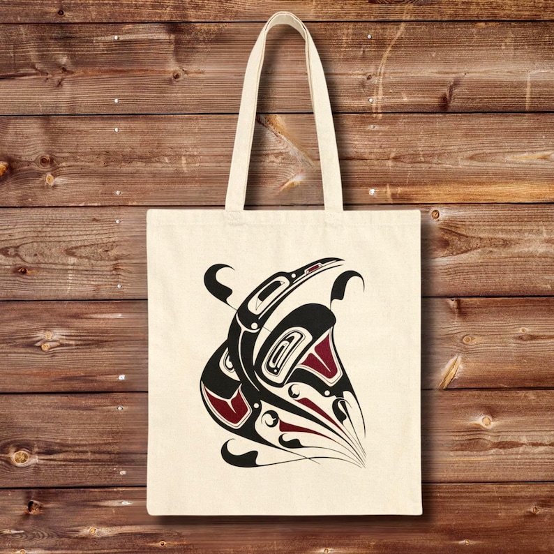 Natural cotton canvas tote bag featuring a bold Northwest Coast–inspired raven totem design in black and deep red, centered on the front. The bag hangs against a warm wooden plank wall background, showing long reinforced shoulder straps and a structured flat-bottom shape.