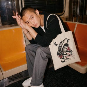Alt text: Young man with a buzz cut sits on an orange subway seat, resting his head on clasped hands, while a cream canvas tote featuring a bold black-and-red orca formline design sits beside him in a moody editorial-style transit scene.