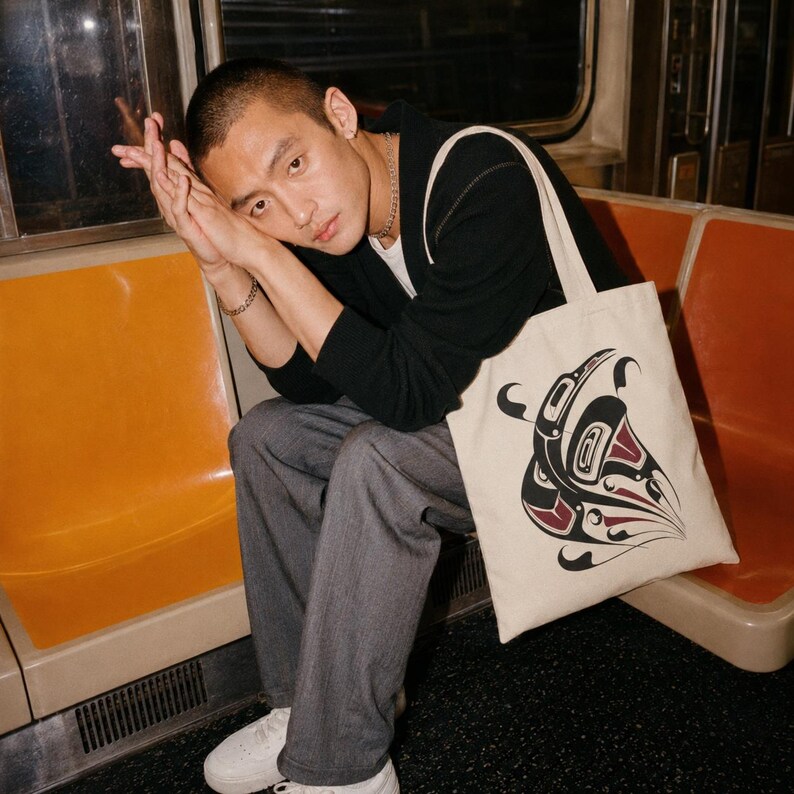 Alt text: Young man with a buzz cut sits on an orange subway seat, resting his head on clasped hands, while a cream canvas tote featuring a bold black-and-red orca formline design sits beside him in a moody editorial-style transit scene.