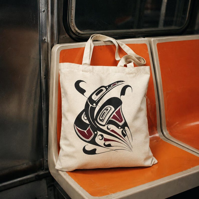 AI-generated lifestyle mockup of a natural cotton canvas tote bag with original Pacific Northwest Coast inspired raven formline artwork, placed upright on an orange NYC subway seat with metal train walls and a moody urban transit vibe.