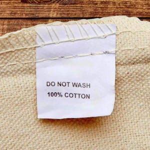 Close-up detail of the inside care label on a cream cotton canvas tote bag, showing reinforced stitching along the seam and a white fabric tag that reads “DO NOT WASH – 100% COTTON.”