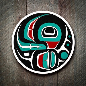May include: Circular sticker featuring a Native American design. The artwork is in black, white, teal, and red, with a stylized bird or animal motif. The sticker is set against a wooden background.