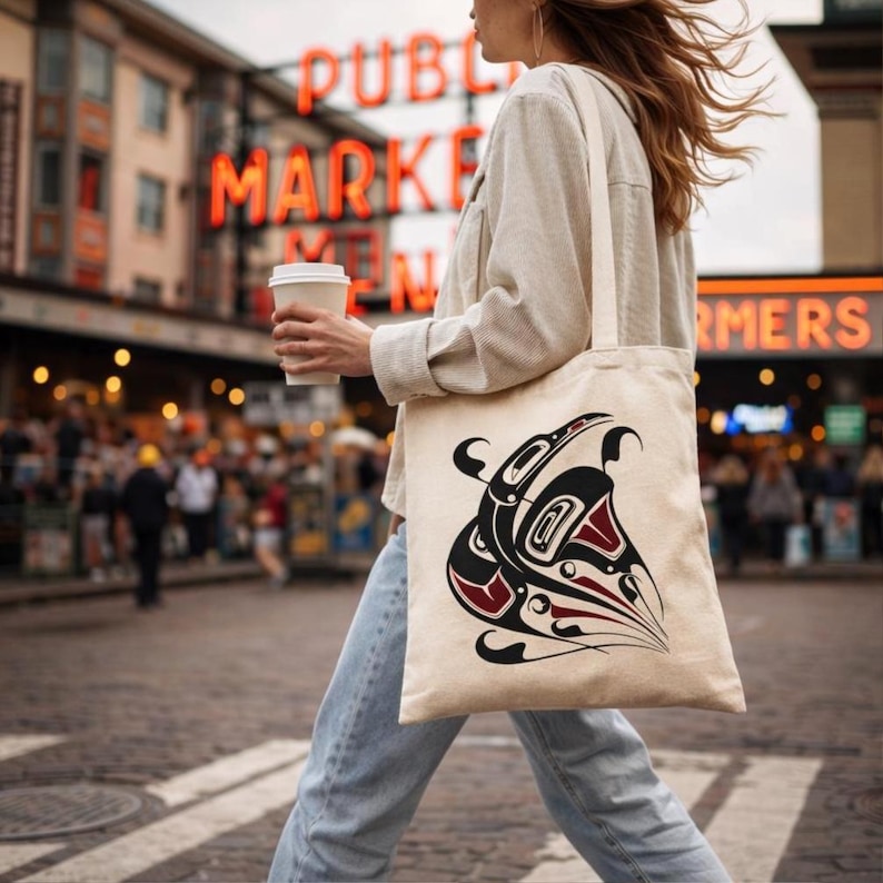Woman walking mid-stride through Pike Place Market in Seattle, holding a white takeaway coffee cup. Wears a cream corduroy jacket and light blue jeans, with her hair blowing in the wind. Over her shoulder is a natural canvas tote bag with matching canvas straps, featuring a bold black and deep red Northwest Coast formline raven totem design. The iconic red “Public Market” neon sign glows softly in the blurred background, with market crowds and warm city lights creating an urban influencer vibe.