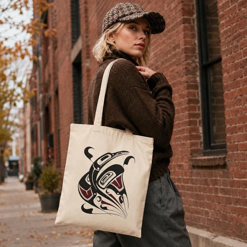 AI-generated lifestyle mockup of a natural cotton canvas tote bag with original Pacific Northwest Coast inspired raven formline artwork, worn by a blonde model in a brown sweater and cap on an autumn brick city street.