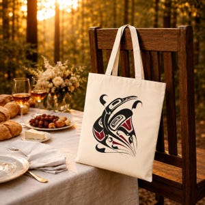 AI-generated lifestyle mockup of a natural cotton canvas tote bag with original Pacific Northwest Coast inspired raven formline artwork, hanging on a wooden chair beside an elegant outdoor autumn picnic table with wine, bread, grapes, flowers, linen, and golden forest light.