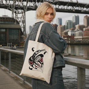 Square lifestyle mockup of a person in casual streetwear wearing a cream cotton tote bag over one shoulder, standing outdoors with a cool Seattle-style urban waterfront backdrop. The tote features a bold Northwest Coast–inspired black, red, and cream orca-style design, shown clearly as the focal point.