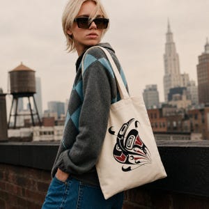 AI-generated lifestyle mockup of a natural cotton canvas tote bag with original Pacific Northwest Coast inspired raven formline artwork, worn over the shoulder by a blonde model in black sunglasses and a gray-blue argyle sweater on a moody NYC rooftop with skyline and water tower in the background.