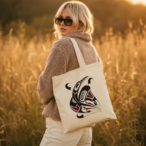 AI-generated lifestyle mockup of a natural cotton canvas tote bag with original Pacific Northwest Coast inspired raven formline artwork, worn over the shoulder by a blonde model in sunglasses and a cozy beige fleece jacket, standing in a golden field at sunset.