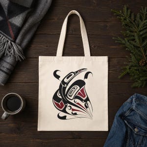 AI-generated top-view flat lay mockup of a natural cotton canvas tote bag with original Pacific Northwest Coast inspired raven formline artwork, styled on a dark wood surface with a gray plaid wool blanket, black coffee, evergreen sprigs, and a folded denim jacket for a minimal Seattle cabin vibe.