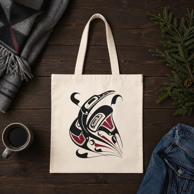 AI-generated top-view flat lay mockup of a natural cotton canvas tote bag with original Pacific Northwest Coast inspired raven formline artwork, styled on a dark wood surface with a gray plaid wool blanket, black coffee, evergreen sprigs, and a folded denim jacket for a minimal Seattle cabin vibe.