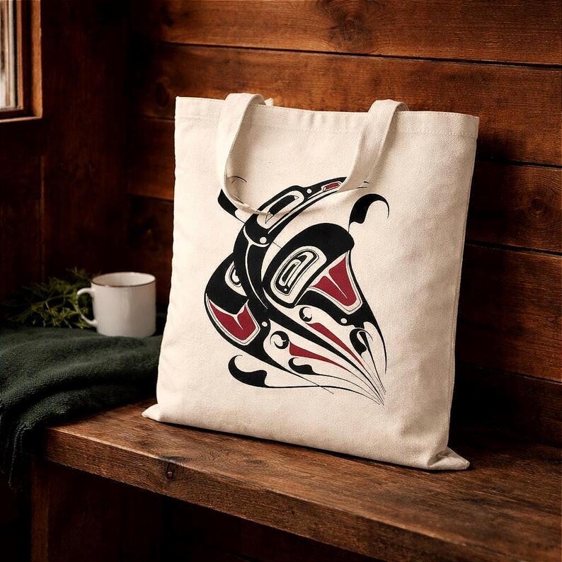 Natural cotton canvas tote bag with bold black and deep red Northwest Coast–inspired raven totem design, displayed on a rustic wooden bench inside a warm cabin setting. Both straps fall to the sides, leaving the full artwork clearly visible against a cedar plank wall background with soft natural light.