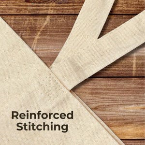 Close-up detail of a natural cotton canvas tote bag handle showing reinforced stitching at the seam. The fabric texture and stitching are clearly visible, highlighting durability and quality construction. The tote is photographed on a turquoise painted wooden background with the text “Reinforced Stitching” displayed in the lower corner.