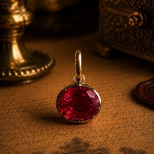 Pode incluir: Pingente de ouro com uma grande pedra preciosa vermelha oval e facetada. O pingente tem uma argola de ouro e uma moldura de ouro ao redor da pedra preciosa. O fundo inclui um castiçal dourado e uma caixa decorativa.