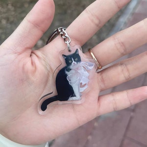 May include: A clear acrylic keychain featuring a black and white cat wearing a white bow. The cat is sitting and looking at the camera.