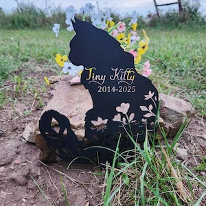 May include: A black metal cat silhouette memorial marker featuring cut-out floral patterns at the base. The text "Tiny Kitty" is printed in a gold script font above the years "2014-2025" in a clean, white, sans-serif typeface.