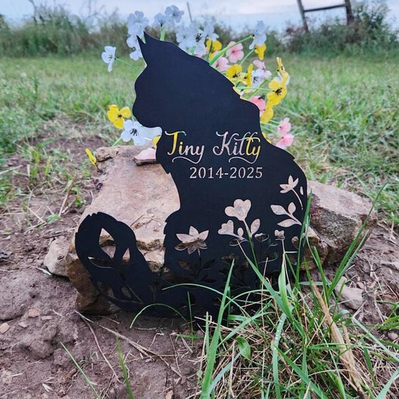 May include: A black metal cat silhouette memorial marker featuring cut-out floral patterns at the base. The text "Tiny Kitty" is printed in a gold script font above the years "2014-2025" in a clean, white, sans-serif typeface.