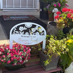 May include: A white metal garden sign featuring a laser-cut floral and butterfly design at the top with the text "Welcome to our Daycare Family" in the center. The sign is positioned on a bench surrounded by various potted plants and flowers.