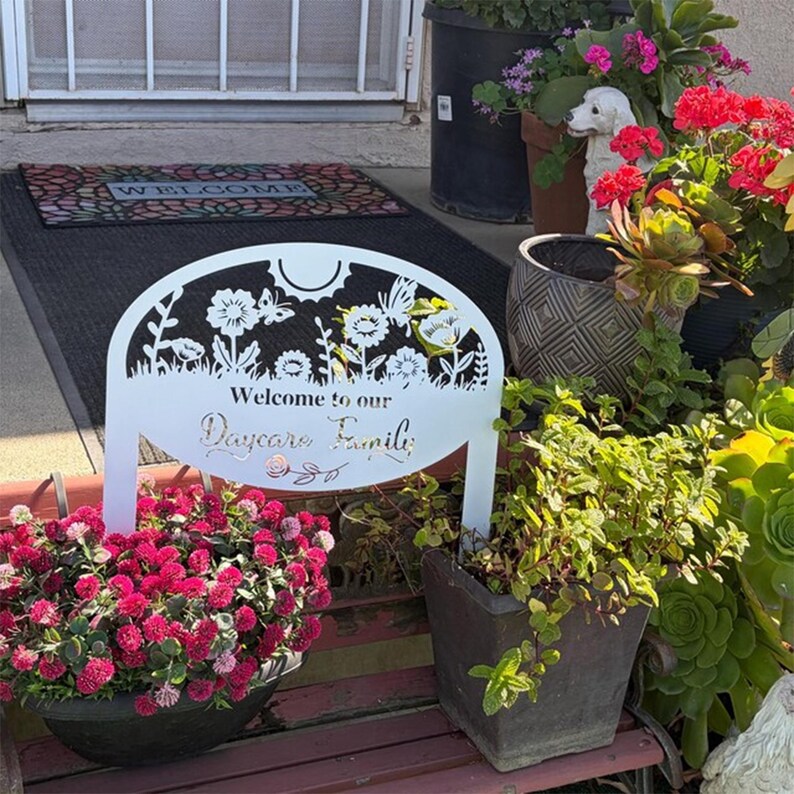 May include: A white metal garden sign featuring a laser-cut floral and butterfly design at the top with the text "Welcome to our Daycare Family" in the center. The sign is positioned on a bench surrounded by various potted plants and flowers.