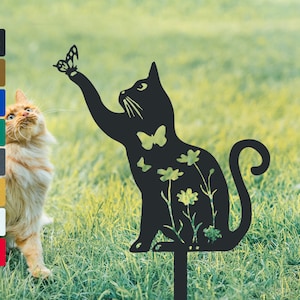 May include: Black metal silhouette of a cat reaching for a butterfly, with flowers and butterflies cut out of the design. The silhouette is on a stake for outdoor garden decor.