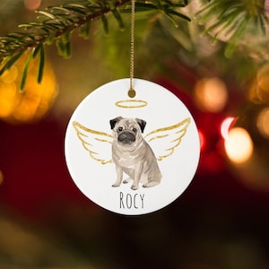May include: A white ceramic Christmas ornament with a pug design, gold angel wings, and a halo. The pug is light brown and black. The name "ROCY" is printed in black. The ornament hangs from a gold string.