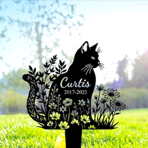 May include: A black metal garden stake featuring a silhouette of a cat surrounded by floral patterns. The name Curtis and the years 2017-2025 are engraved in white script and sans-serif font on the body of the cat, which is perched on a stake.