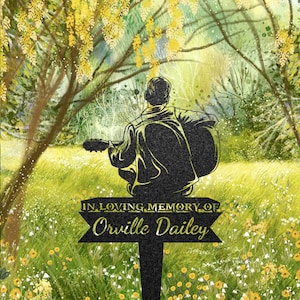 Könnte beinhalten: Eine schwarze Metallsilhouette einer Person, die Gitarre spielt, in einem Feld mit gelben und weißen Blumen. Die Silhouette ist an einem schwarzen Metallpfahl befestigt. Ein Schild lautet "In liebevoller Erinnerung an Orville Dailey".