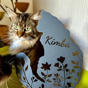 May include: A dark gray metal silhouette of a cat with a long, fluffy tail, featuring the name Kimba laser-cut into the center and intricate floral cutouts along the lower body. The cat-shaped plaque has a textured finish and stands upright.