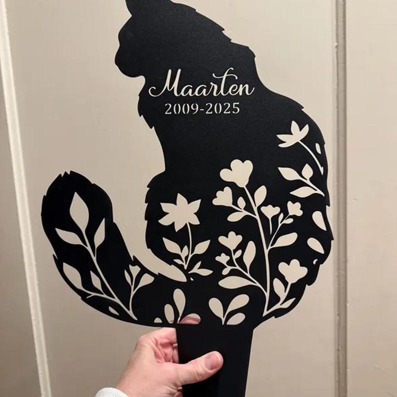 May include: A black metal garden stake shaped like a sitting cat with a long, curved tail. The silhouette features cut-out floral patterns and the white text "Maarten" followed by the years "2009-2025" printed on the cat's upper body.