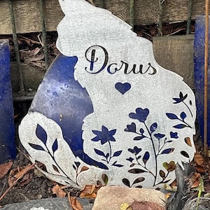 May include: A silver-colored metal garden stake shaped like a cat silhouette. The cutout design features the name "Dorus" in cursive script, a small heart, and various floral and leaf patterns throughout the body of the cat figure.