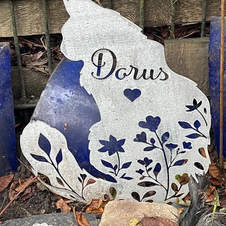 May include: A silver-colored metal garden stake shaped like a cat silhouette. The cutout design features the name "Dorus" in cursive script, a small heart, and various floral and leaf patterns throughout the body of the cat figure.