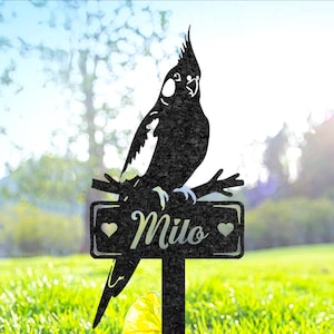 以下が含まれることがあります： 黒い金属製の庭の杭。オウムのシルエットが枝にとまり、その上に看板があります。「Milo」という名前が筆記体で表示され、ハートのシンボルが付いています。庭での屋外使用に適しています。