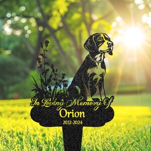 以下が含まれることがあります： 犬のシルエット、草、そして「In Loving Memory Of Orion 2012-2024」のテキストが特徴の黒い金属製の記念杭。犬は金色の首輪と胸のマーキングをしています。背景は、ぼやけた緑のフィールドと太陽光です。