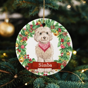 May include: A white ceramic Christmas ornament featuring a watercolor illustration of a small, white dog with a red bandana, sitting in a wreath of green holly, red berries, and gold ornaments. The ornament has the name "Simba" written on a red ribbon.