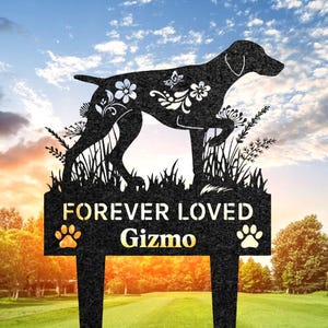 May include: Black metal garden stake featuring a dog silhouette with floral and butterfly accents. The stake reads "FOREVER LOVED" and "Gizmo" with paw prints. The background shows a sunset and trees.