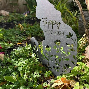 May include: A silver-toned metal garden stake shaped like a sitting cat with a glittery finish. The silhouette features cut-out floral patterns and is engraved with the name Tippy and the years 2007-2024 in a dark, elegant script and font.