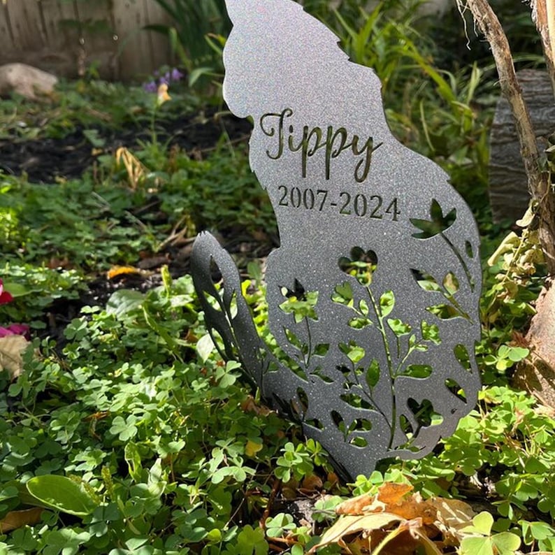 May include: A silver-toned metal garden stake shaped like a sitting cat with a glittery finish. The silhouette features cut-out floral patterns and is engraved with the name Tippy and the years 2007-2024 in a dark, elegant script and font.