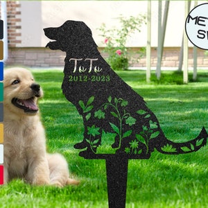 Könnte beinhalten: Schwarze Metallsilhouette eines Hundes mit Blumen und dem Text "Toto 2012-2023". Die Silhouette befindet sich auf einem schwarzen Metallpfahl. Ein Golden Retriever-Welpe sitzt im Gras vor der Silhouette.