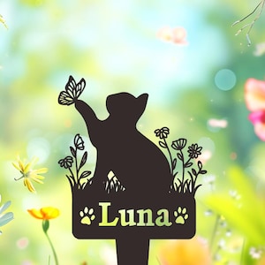 May include: A black metal garden stake featuring a silhouette of a cat reaching toward a butterfly, surrounded by floral stems. The rectangular base displays the name Luna in a serif font, flanked by two small paw print icons on each side.