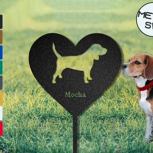 Puede incluir: Estaca de jardín de metal negro en forma de corazón con una silueta de un perro beagle dentro del corazón. La estaca tiene la palabra "Mocha" escrita debajo del perro. Un perro beagle está de pie en la hierba junto a la estaca.