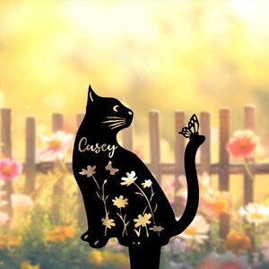 May include: A black metal garden stake shaped like a sitting cat with a butterfly perched on its tail. The cat silhouette features cut-out floral patterns and the name Casey written in a script font on the side of the cat's body.
