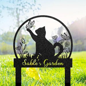 May include: Black metal garden stake featuring a silhouette of a cat reaching for a butterfly within a circular frame. The words "Sable's Garden" are displayed below the cat. The design includes floral and butterfly cutouts.