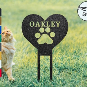 Op de afbeelding: Zwarte metalen hartvormige tuinpaal met een pootprint en de naam "OAKLEY" in gouden letters. De paal staat in een grasachtig gebied.
