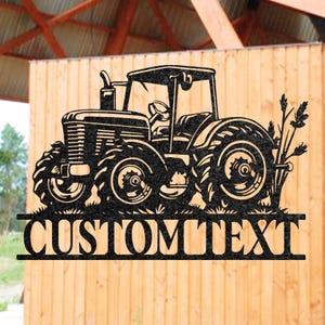 Puede incluir: Silueta de tractor de metal negro con el texto "CUSTOM TEXT" en la parte inferior. El tractor tiene ruedas grandes y un diseño detallado, con espigas de trigo en el lado derecho. El letrero está montado en una pared de madera.