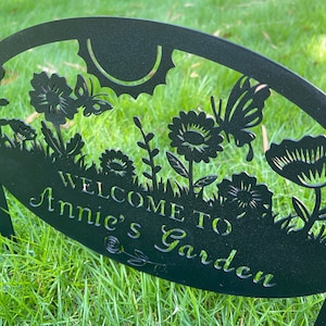 May include: A black metal garden sign with a cut-out design featuring flowers, butterflies, and a sun. The text "WELCOME TO Annie's Garden" is prominently displayed in the center of the oval-shaped plaque, which is supported by two metal stakes.