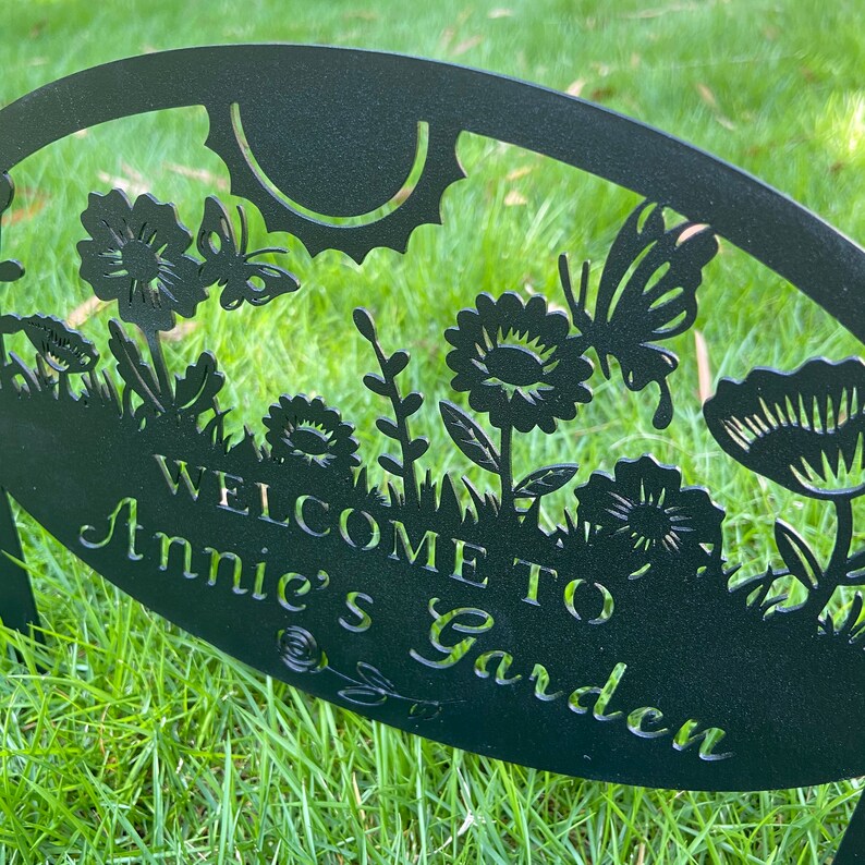 May include: A black metal garden sign with a cut-out design featuring flowers, butterflies, and a sun. The text "WELCOME TO Annie's Garden" is prominently displayed in the center of the oval-shaped plaque, which is supported by two metal stakes.