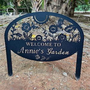May include: A black metal garden sign with a semi-circular top featuring cutouts of flowers, butterflies, and a sun. The text "WELCOME TO Annie's Garden" is centered on the sign, with a small rose illustration below the text and two stakes.