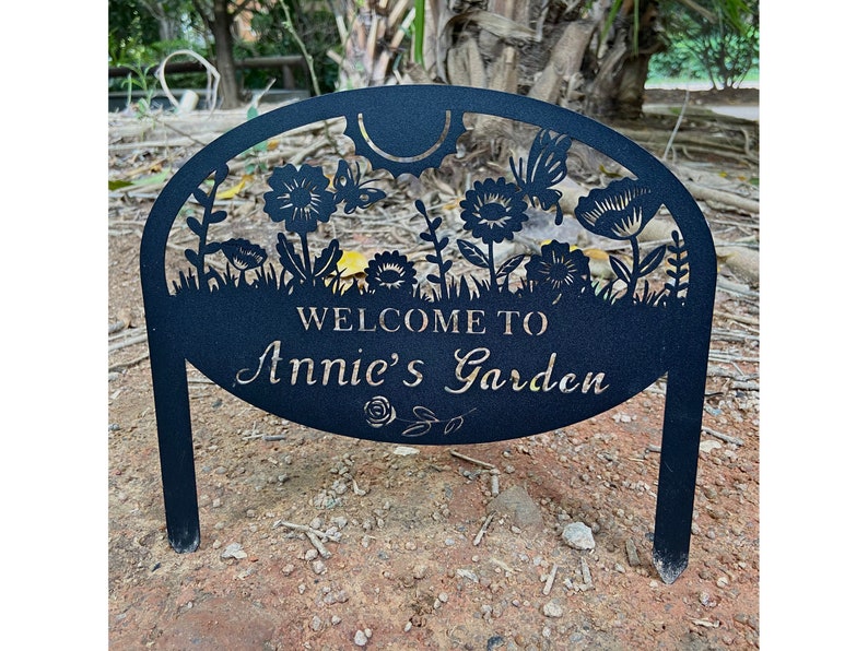 May include: A black metal garden sign with a semi-circular top featuring cutouts of flowers, butterflies, and a sun. The text "WELCOME TO Annie's Garden" is centered on the sign, with a small rose illustration below the text and two stakes.